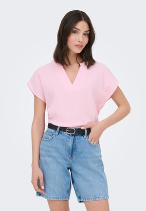 Woman with shoulder-length dark hair wearing a light pink V-neck top tucked into belted light blue denim shorts.