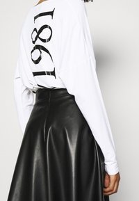 Person in einem weißen Langarmshirt mit "1981" in Schwarz auf dem Rücken und einem hoch sitzenden schwarzen Lederrock vor einem schlichten Hintergrund.