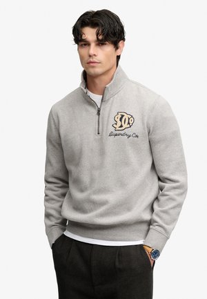 Junger Mann mit dunklen Haaren, der einen hellgrauen Quarter-Zip-Sweatshirt mit Superdry Co. Logo und dunkle Hosen trägt, posiert mit den Händen in den Taschen.