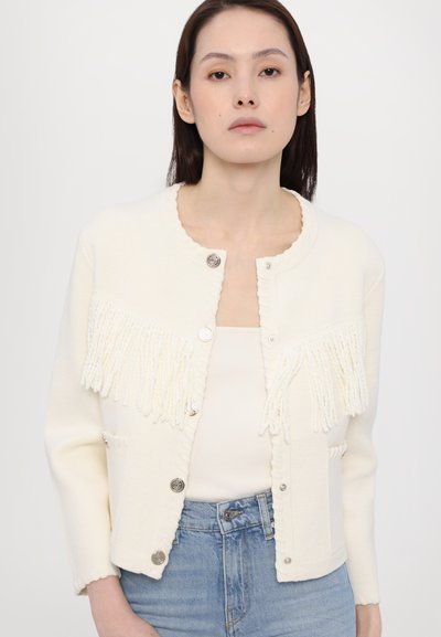Cremfarbener Strickcardigan mit Fransen, rundem Ausschnitt und silbernen Knöpfen. Getragen über einem eng anliegenden beigen Oberteil und hellblauen Jeans.
