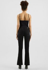 Schwarze Schlagjeans mit tailliertem Bund und zwei Gesäßtaschen, kombiniert mit einem schwarzen Spaghetti-Träger-Oberteil. Glatter Stoff mit subtiler satinierter Oberfläche.