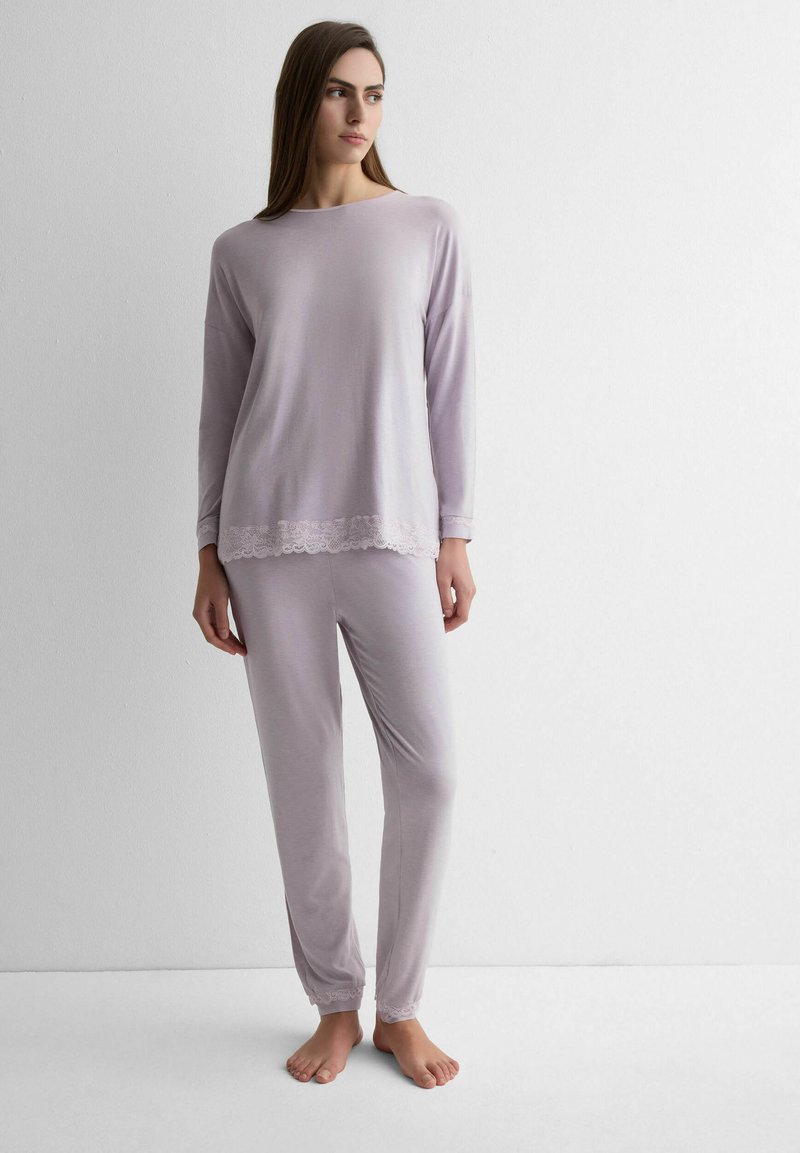 Frau steht barfuß, trägt ein helllila langärmliges Oberteil und passende Hosen mit Spitzenbesatz an Bündchen und Saum, vor einer weißen Wand.