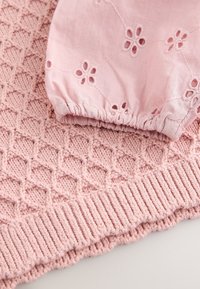 Lyserødt strikket stof med et diamantmønster, der har en matchende lyserød ærme med blomsterbroderi og elastik i samlingen ved manchet.