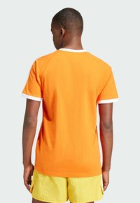 Camiseta de algodón naranja con cuello redondo y puños de manga en blanco, con un diseño simple y mangas cortas. Combinada con pantalones cortos amarillos.