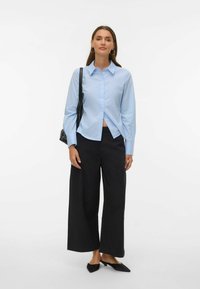 Camisa de botones azul claro de manga larga y cuello, combinada con pantalones anchos negros y mules negros de tacón. Complemento de bolso negro.