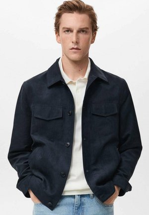 Chaqueta de ante azul marino con cierre de botones, cuello puntiagudo y dos bolsillos en el pecho. Llevada sobre una camiseta polo blanca y unos jeans azules. Textura suave.