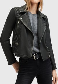 Giacca da biker in pelle nera con spalle trapuntate, zip asimmetrica, due tasche frontali con zip e design cropped. Indossata sopra un maglione grigio.