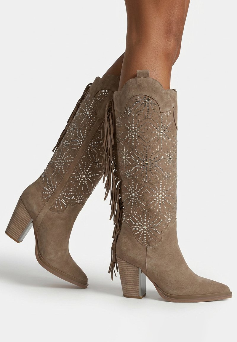 Bottes en daim taupe montant jusqu’aux genoux avec talons carrés, motifs en strass en forme d’étoile et détails de franges latérales, portées jambes croisées.