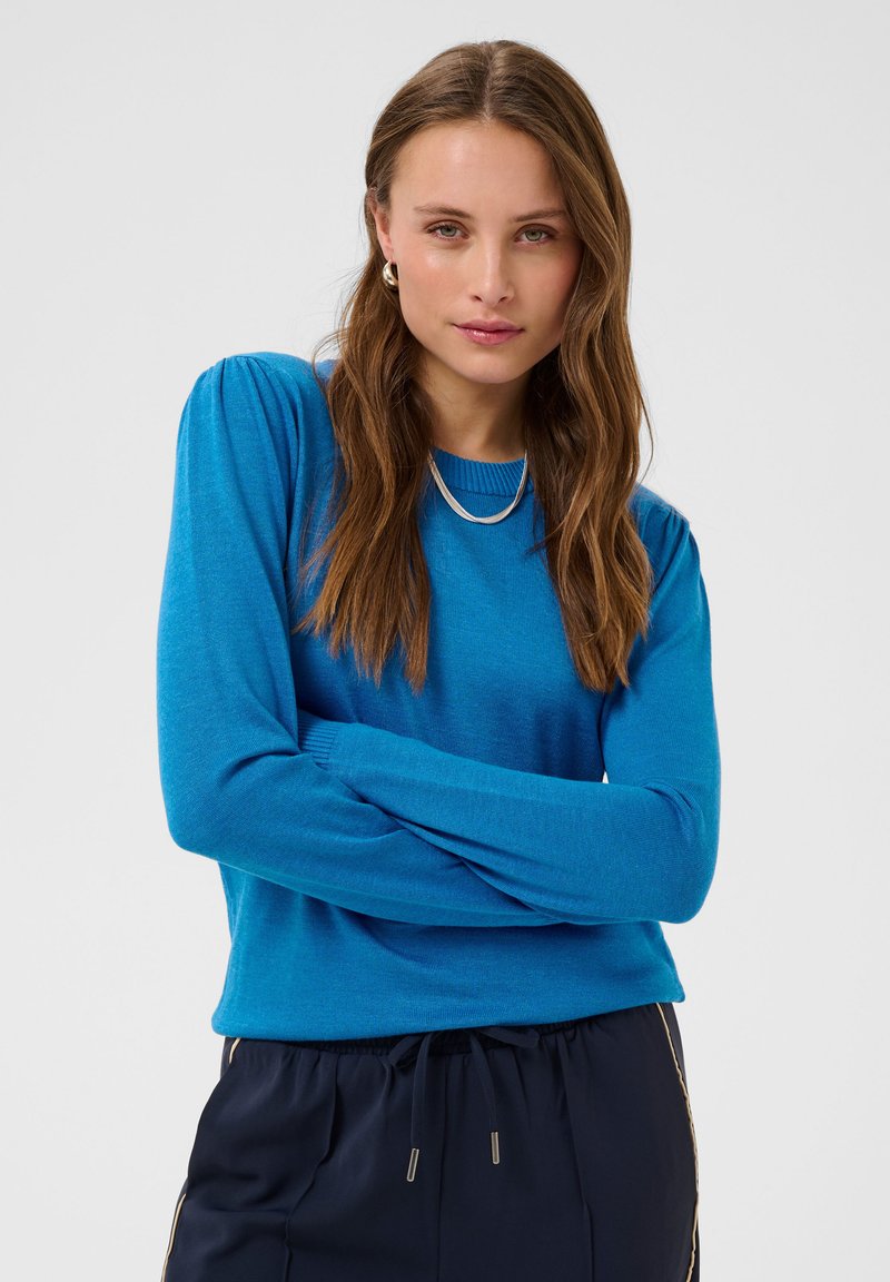 Jeune femme aux longs cheveux bruns portant un pull bleu vif et un pantalon noir, debout les bras croisés devant un fond uni.