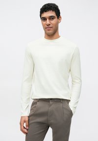 Langärmliges, cremefarbenes Baumwoll-T-Shirt mit Rundhalsausschnitt, kombiniert mit braunen Hosen. Eng anliegender Stil mit glatter Textur.