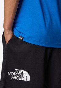 Blå kortärmad skjorta med en slät textur, tillsammans med svarta shorts med en vit logotyp. Etiketter synliga på skjortan och shortsen.