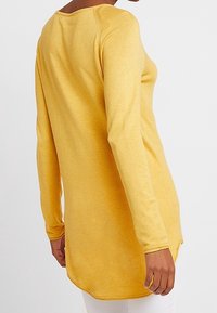 Tunique jaune à manches longues avec un col rond et des fentes latérales. Texture douce et ourlet arrière allongé pour une silhouette décontractée.