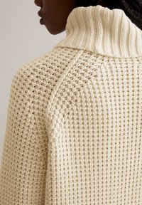 Cremfarbener Strickpullover mit hohem, geripptem Kragen, strukturiertem Gewebe und lockerer, entspannter Passform mit weiter Ausschnitt.
