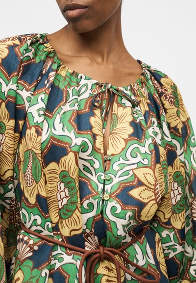 Γυναίκα που φοράει πολύχρωμο φόρεμα με λουλούδια σε πράσινα, κίτρινα και καφέ σχέδια, με κουμπιά και δεμένη ζώνη στη μέση.