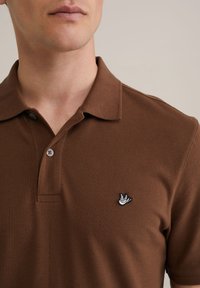 Polo marrón con una tela texturizada, dos botones en el cuello y un pequeño logo bordado de un pájaro blanco en el lado izquierdo del pecho.
