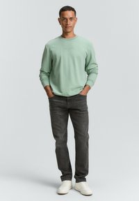 Felpa verde menta con polsini a costine, abbinata a jeans grigi scuri e sneakers bianche. Il modello sta in piedi con le mani in tasca davanti a uno sfondo neutro.