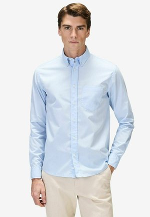 Mand iført lyseblå skjorte med knapper og forlomme samt beige bukser, stående med den ene hånd i lommen.