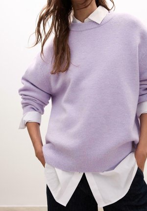 Femme portant un pull en tricot violet clair sur une chemise blanche à col, manches retroussées, mains dans les poches, sur un fond neutre.