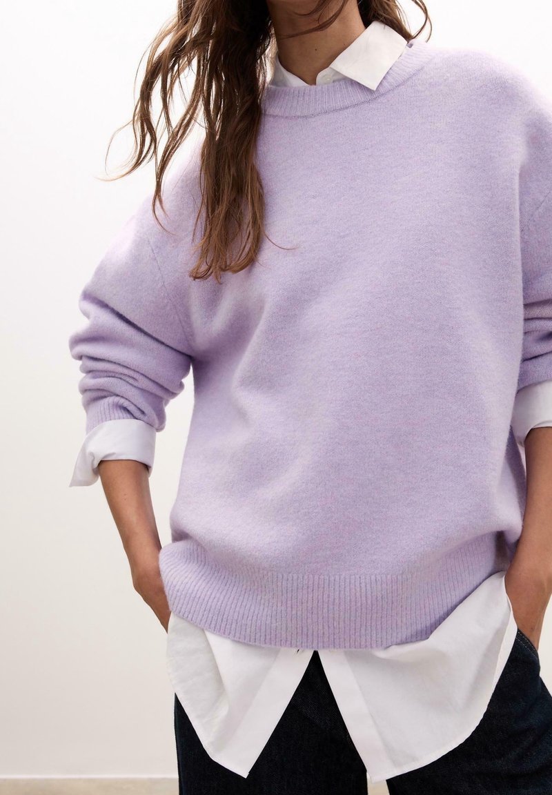 Femme portant un pull en tricot violet clair sur une chemise blanche à col, manches retroussées, mains dans les poches, sur un fond neutre.