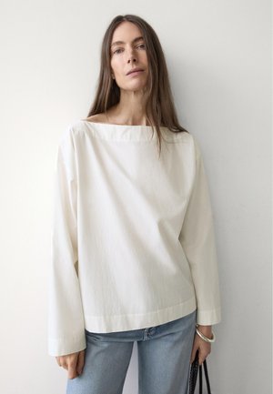 Femme aux longs cheveux bruns portant un haut blanc ample à manches longues et un jean bleu clair, tenant un sac noir texturé à ses côtés.