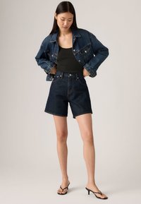 Σκούρο τζιν μπουφάν με ανοιχτή ραφή, σε συνδυασμό με μαύρο αμάνικο τοπ και σκούρα τζιν σορτς. Το μοντέλο φοράει μαύρα σανδάλια με τακούνι.