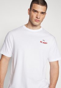 Balts kokvilnas T-krekls ar apaļu kakla izgriezumu un īsām piedurknēm, ar sarkanu izšūtu "ST. PAULI" logo un Puma logo uz krūtīm.