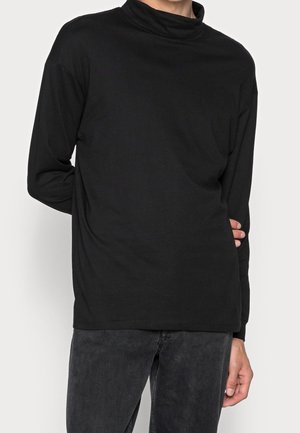 Person trägt ein schwarzes Langarmshirt mit Rollkragen und dunkelgraue Jeans, mit einem Arm hinter dem Rücken angewinkelt.