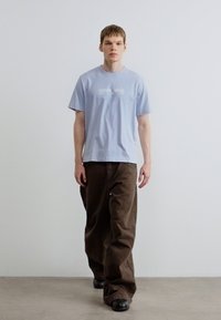 Svetlomodré bavlnené tričko s bielym logom, v kombinácii s voľnými hnedými nohavicami. Čierna obuv dopĺňa outfit. Jednoduché pozadie.