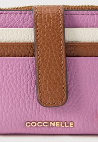 Close-up of a textured leather wallet with pink, white, and brown sections, featuring a brown leather strap and gold "COCCINELLE" logo.