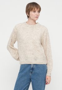 Rövid barna hajú nő, aki texturált bézs kötött pulóvert és kék farmernadrágot visel, oldalra néz.