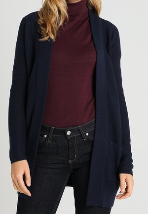 Femme portant un cardigan bleu marine ouvert sur un col roulé bordeaux et un jean noir, debout, les mains détendues le long du corps.