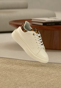 Λευκό αθλητικό παπούτσι με λεπτομέρεια λεοπάρ στο τακούνι και τη γλώσσα, λευκά κορδόνια, παχύ πέλμα, να αιωρείται πάνω από μπεζ χαλί σε περιβάλλον καθιστικού.