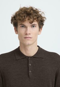 Barna pólóing gallérral, három gombos gombolással és puha textúrával, egy göndör hajú személyen viselve, semleges háttérrel.