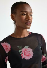 Sort gennemsigtig top med pink rosetryk, med en rund halsudskæring. Accessoriseret med sølv statement øreringe med hængende krystaller.