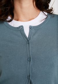 Licht turquoise vest met een geribbelde structuur, ronde halslijn en vijf bijpassende knopen aan de voorkant, gedragen over een wit overhemd.