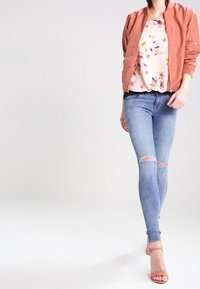 Růžová bomber bunda přes květinovou blůzu, skombinovaná se světle modrými skinny džíny s roztržením na kolenou a růžovými sandály na podpatku.