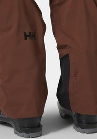 Waterdichte bruine buitenbroeken met zwarte accenten, met HELLY TECH branding, en robuuste zwarte laarzen met gestructureerde zolen.
