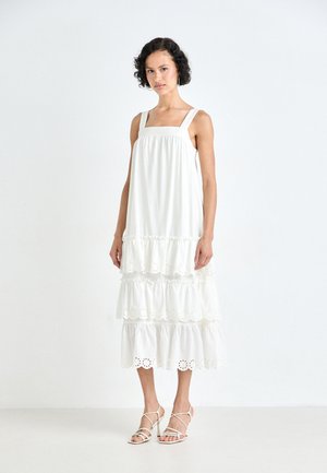 Robe blanche à volants avec décolleté carré, ourlet froncé et détails en broderie anglaise. Le tissu semble doux et fluide. Le modèle porte des sandales à lanières.