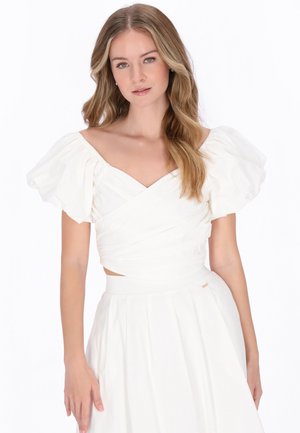 Kobieta o długich blond włosach, ubrana w biały top z odsłoniętymi ramionami i bufiastymi rękawami oraz dopasowaną białą spódnicę na jednolitym tle.