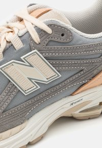 Grå og beige treningssko med et teksturert stoffoverdel, detaljert søm og en lys farget gummisåle. Har et fremtredende "N"-logo.