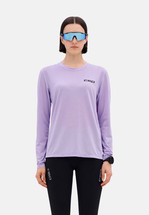 Woman wearing reflective blue sports sunglasses, a lavender long-sleeve shirt with logo, black leggings, and a black smartwatch on left wrist.