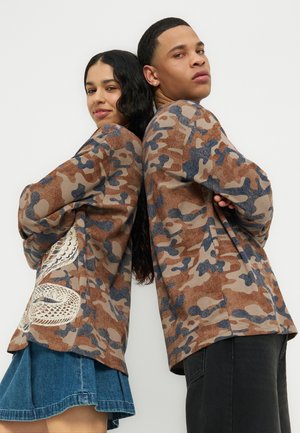 Deux personnes se tiennent dos à dos, les bras croisés, portant des vestes assorties à motifs camouflage marron et bleu, dont l'une arbore un motif de serpent blanc.