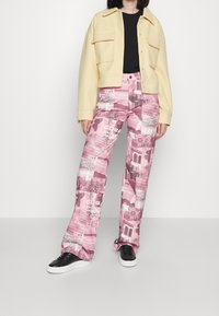 Un pantalon avec un design graphique rose comportant des motifs floraux et du texte, associé à une veste courte jaune et des baskets noires.