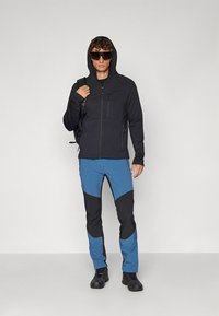 Sort jakke med lynlås og hætte, kombineret med blå og sorte mønstrede bukser. Modellen bærer også mørke solbriller og en rygsæk.