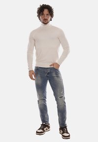 Maglione bianco a collo alto abbinato a jeans blu sbiaditi. Sneakers marroni con dettagli bianchi. Tessuto morbido, vestibilità slim, leggero invecchiamento sui jeans.