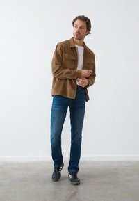 Hombre ajustando la manga de una chaqueta de pana marrón, lleva una camiseta blanca, jeans azules y zapatillas negras, de pie sobre un suelo gris contra una pared blanca.