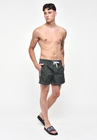 Costumi da bagno verdi con cintura regolabile, tasche laterali e strisce gialle a contrasto. Indossati con ciabatte nere con logo bianco.