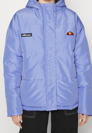 Personne portant une doudoune violet clair avec capuche, patch logo sur la poitrine et pantalon noir avec coutures blanches.