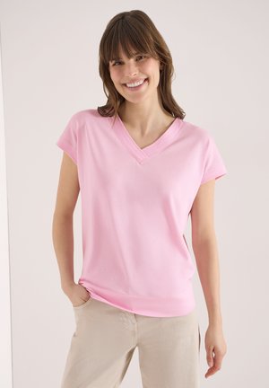 Femme souriante portant un t-shirt à manches courtes rose pâle avec un col en V et un pantalon beige, une main dans la poche, sur un fond uni.