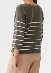 Osoba ubrana w oliwkowozielony sweter z białymi poziomymi paskami i beżowe spodnie, widziana od tyłu i z boku na jednolitym tle.